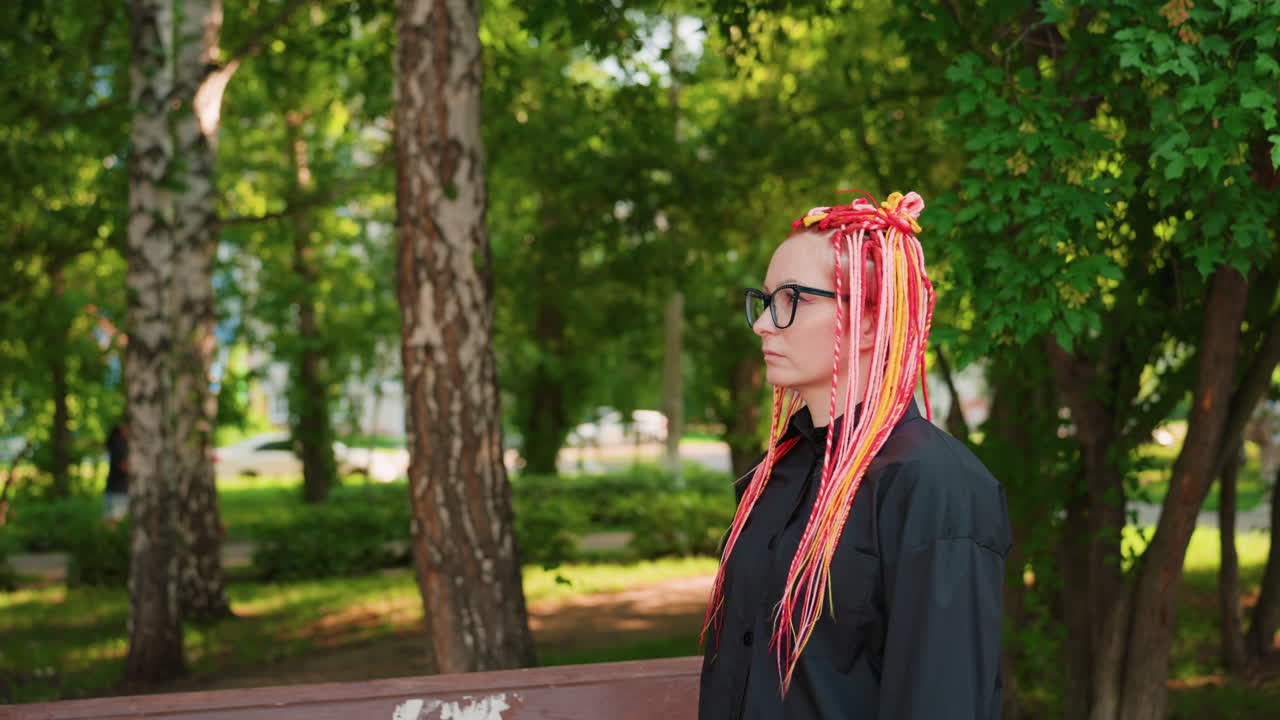 어두운 코트와 안경을 착용하고 나뭇잎이 덮인 길과 자작나무 줄기가 보이는 그늘진 공원에서 나무 벤치를 지나 걸어가는 네온 브레이드 머리를 한 백인 학생, 시선은 아래로 향하고 느린 걸음, 수업 후 사색에 잠긴 분위기