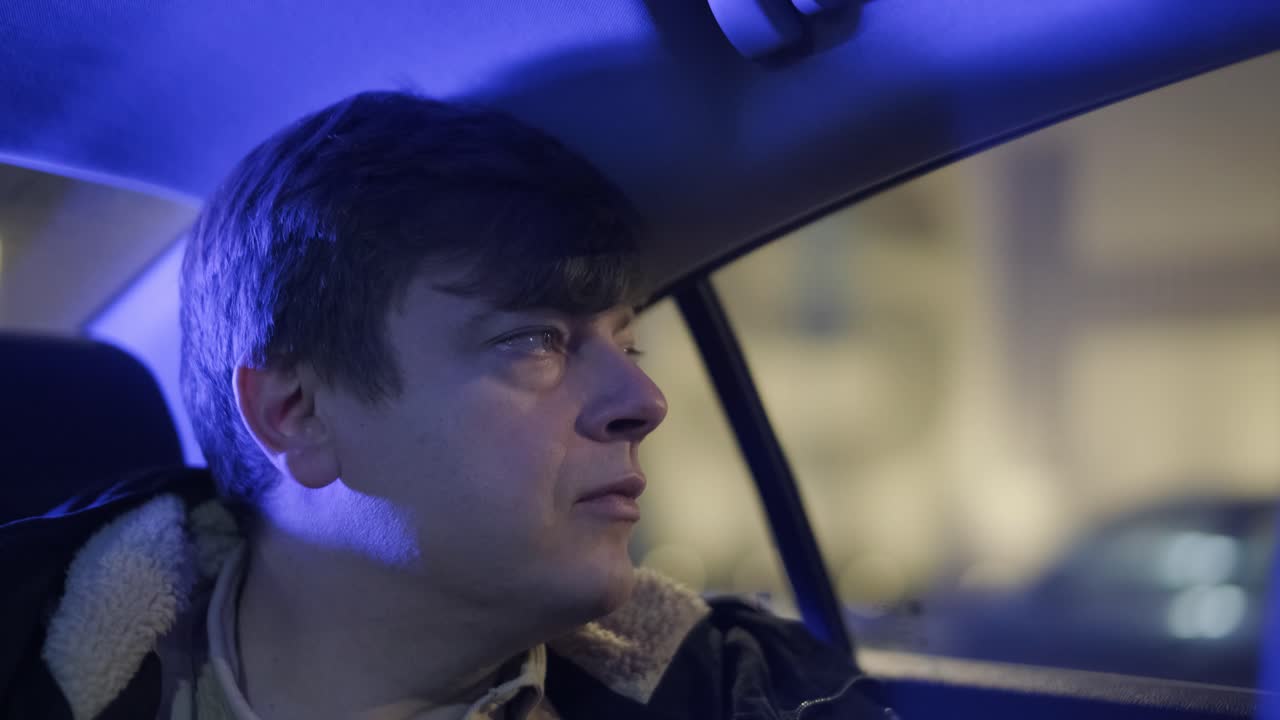 멋진 젊은 사업가의 느린 동작 클로즈업은 밤에 도시의 중심에서 운전자와 함께 현대적인 차로 여행하는 동안 밖을 바라보고 있습니다.