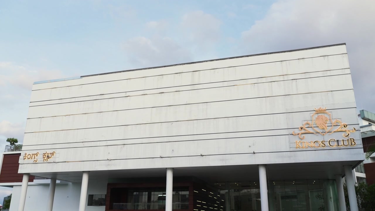 Ripresa esterna con panoramica verso il basso della facciata di un lussuoso club sportivo indiano sotto un cielo serale tenue, adatta come inquadratura di apertura per qualsiasi evento.