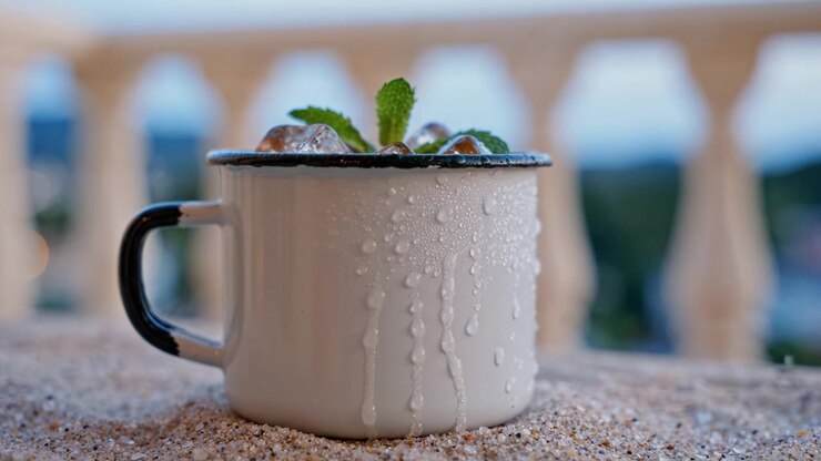 Refreshing Minty Drink Served in an Enamel Mug with Ice Cubes on a Sandy Surface Near a White Railing Overlooking a Scenic Outdoor View at Dusk