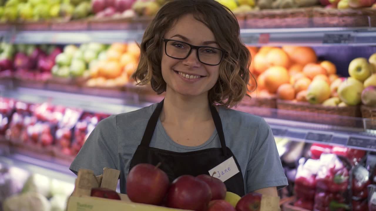 Beautiful smiling young female supermarket employee in black apron holding a box full of apples in front of shelf in supermarket with pretty face looking at camera professional front portrait startup business. Slow motion