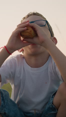 una mujer con un vestido azul sostiene dos bocadillos mientras ofrece uno a su hijo pequeño, que lleva gafas y una camisa blanca. están sentados juntos en un campo cubierto de hierba, disfrutando de un momento pacífico al aire libre