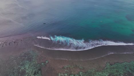 toma aérea de un avión no tripulado de gran ángulo de un barco de pesca indonesio local que navega por la costa y el arrecife de coral, con surfistas remando y olas rodando hacia la costa en bali