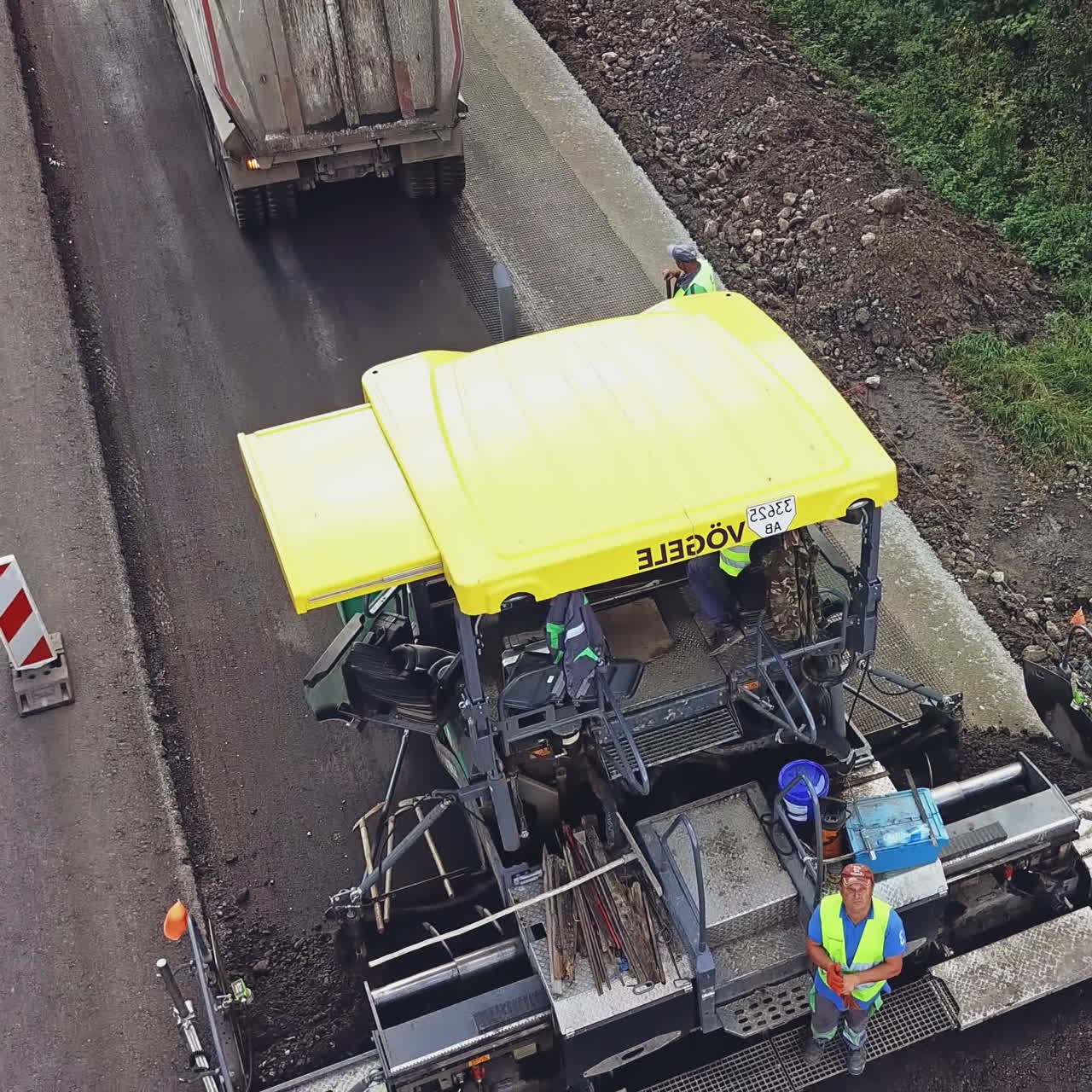 A truck with bitumen slowly drives up to a paver on a highway near a forest landing on the background of road repair works near the city. Aerial view.