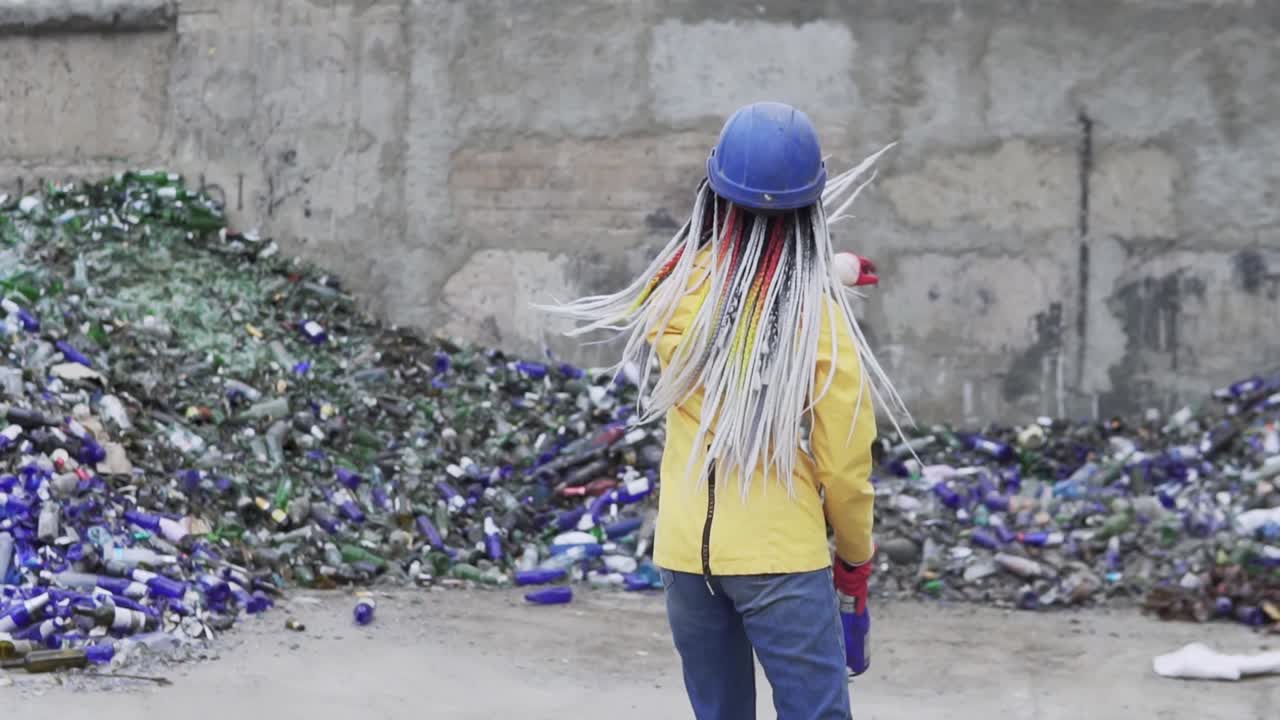 mujer con sombrero duro de pie contra la pila de vidrio roto, botellas usadas junto a la pared. niña con chaqueta amarilla rompiendo viejas botellas de vidrio para su reciclaje. vista rara. cámara lenta