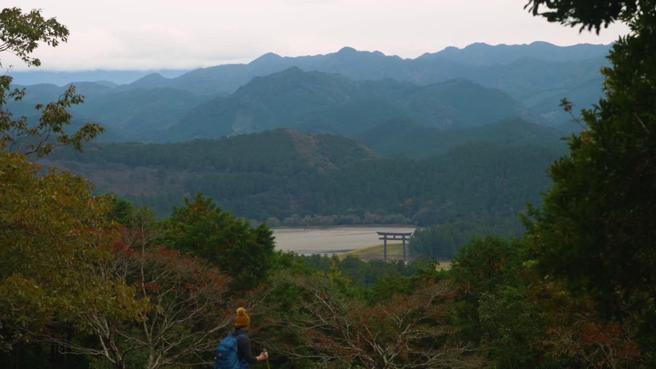 excursionista caminando por el paisaje de montañas boscosas y la lejana puerta torii, kumano kodo
