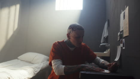 tattooed inmate writing letter at desk in prison cell