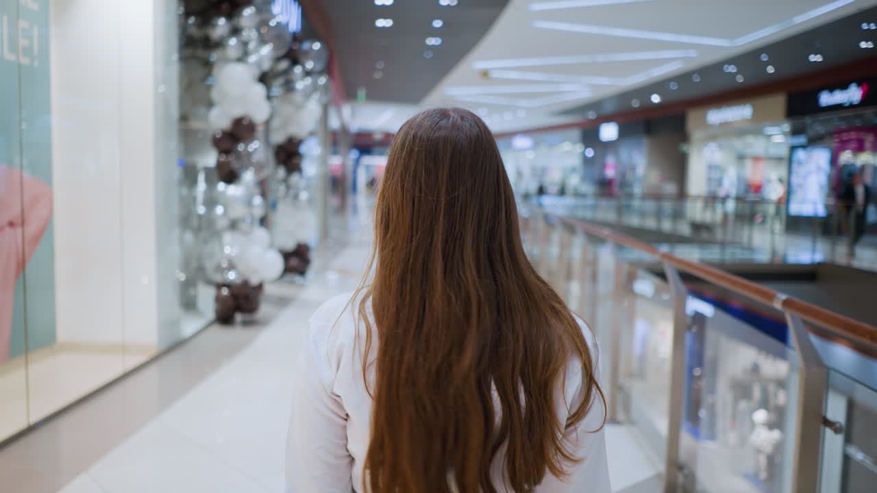 화려한 쇼핑백을 들고 현대 쇼핑몰을 걸어다니는 안경을 입은 자신감 넘치는 여성의 뒷면, 그녀는 밝은 상점 불빛으로 비는 소매 공간에서 의도적으로 걸어갑니다.