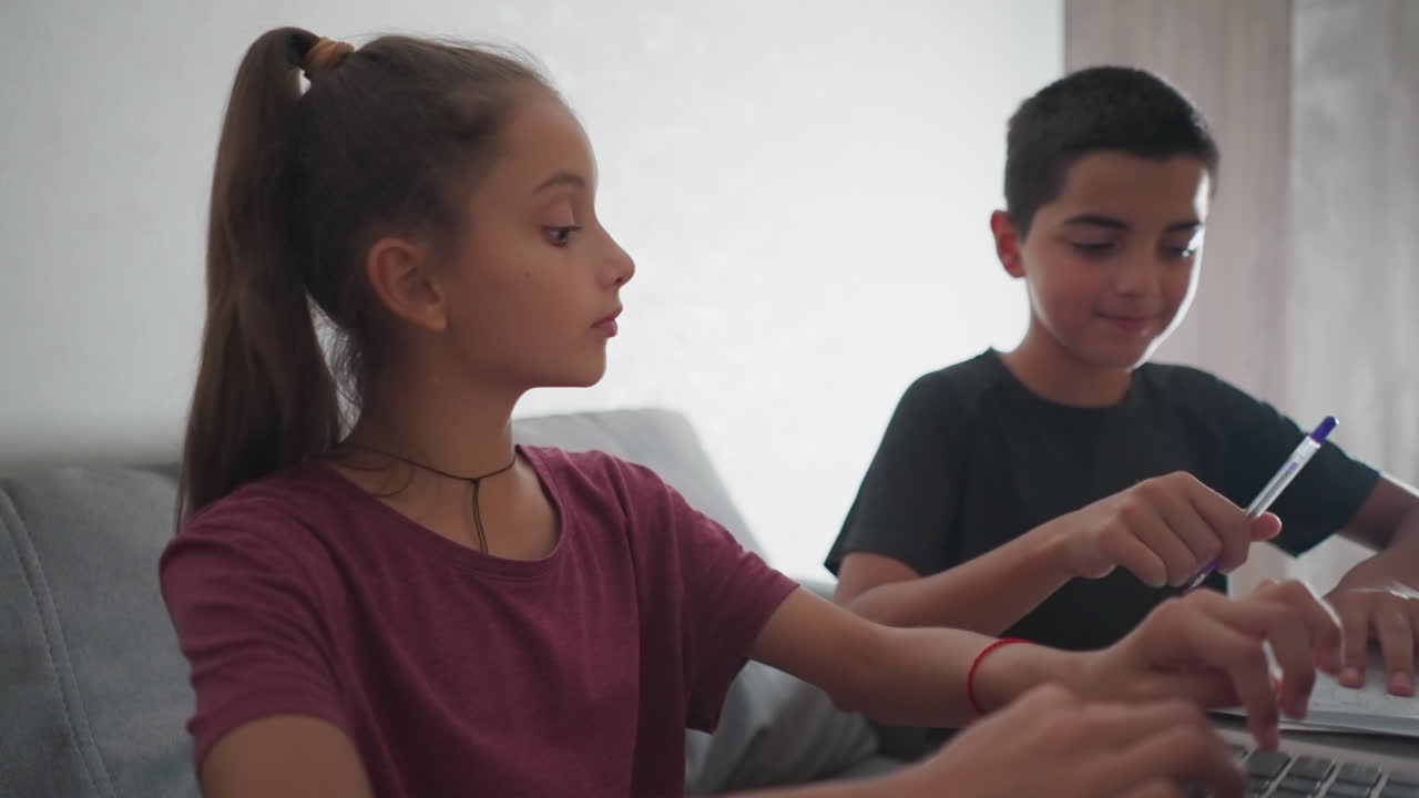 close up of boy writing with pen taps girl wearing red rope bracelet as she types on laptop she pauses looks at his work smiles gives thumbs up while boy laughs happily and continues writing focused