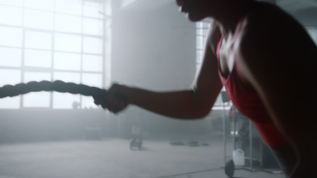 Deportista afroamericana usando cuerdas de batalla durante su entrenamiento en el gimnasio.