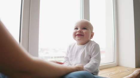 mother and her baby son having fun and playing at home. little kid 1 years old play with his mom arms at home near a big window