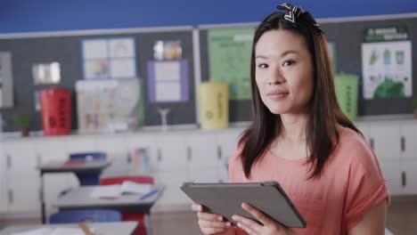 Portrait-of-happy-asian-female-teacher-with-tablet-in-elementary-school-classroom,-slow-motion