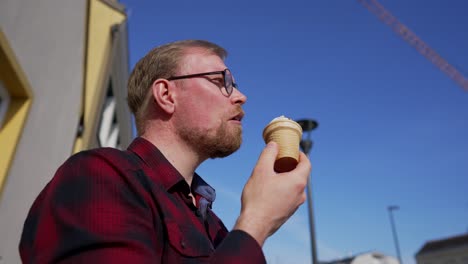 young man with beard and glasses eats ice cream outside on sunny day, low angle