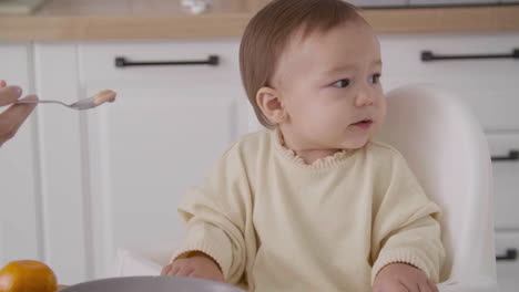 Cute-Little-Girl-Sitting-In-High-Chair-In-The-Kitchen-While-Her-Unrecognizable-Mom-Feeding-Her-1