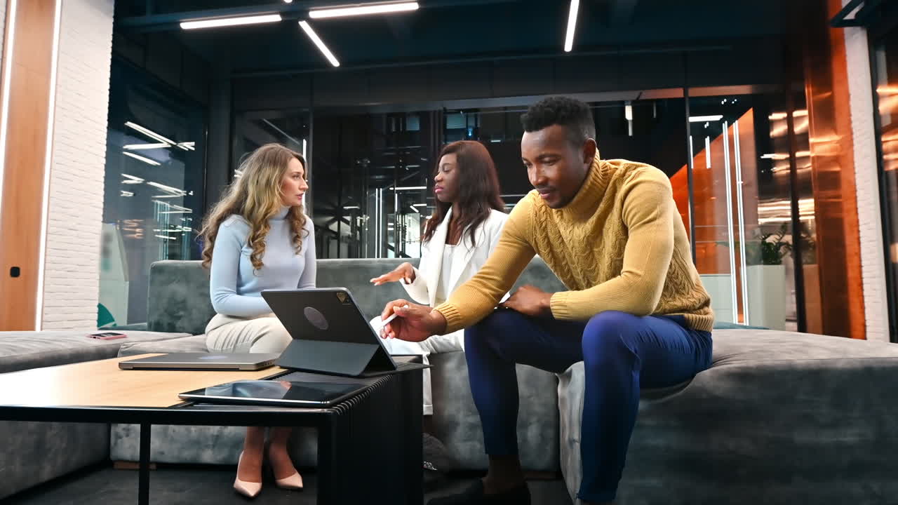 Caucasian and black female workers discussing business while a black man is using a tablet, sitting on a sofa in an office. Slow motion