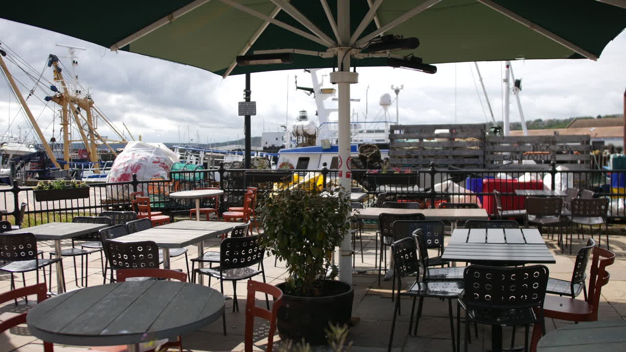 Outdoor café tables and chairs sitting under a large green umbrella near the harbor, with moored boats and fishing gear visible in the background