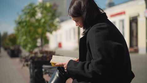 close-up of woman reading outdoors on a sunny day, her hair and greenery in the background gently swaying in the wind, blurred passerby adds motion to the tranquil urban setting