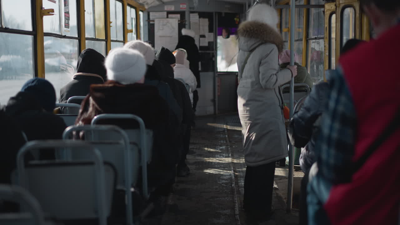 crowded train interior with winter commuters seated while one rider stands gripping pole as carriage slows toward stop, cold light through windows, metal frames adding gritty urban transit mood