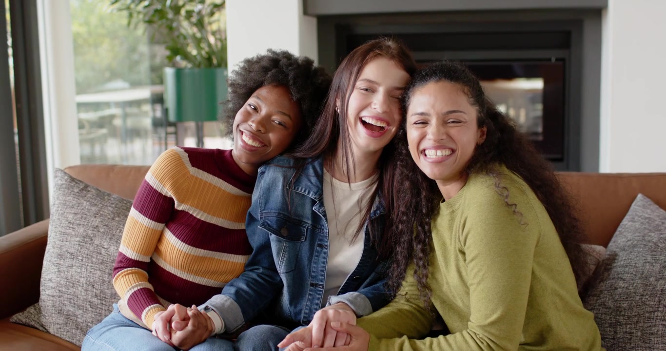 Amigas diversas rindo depois de compartilhar uma piada, encostadas e de mãos dadas no sofá perto da lareira