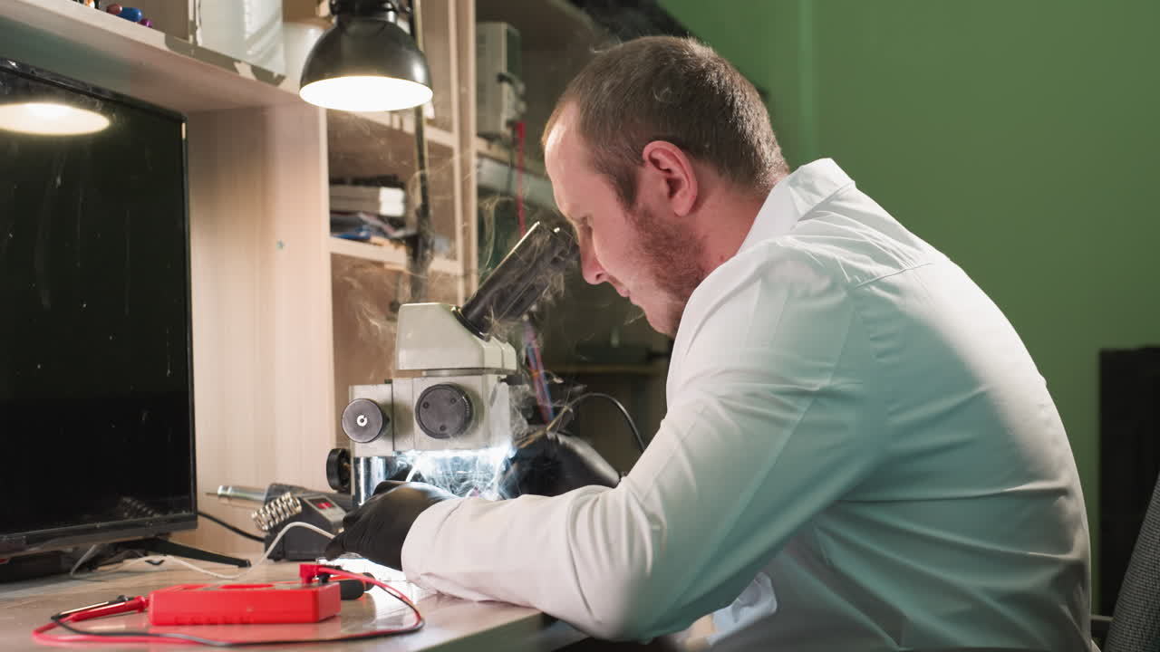 A close-up view of a technician in a white lab coat working on a circuit board under a microscope in his workshop, with other equipment on the table