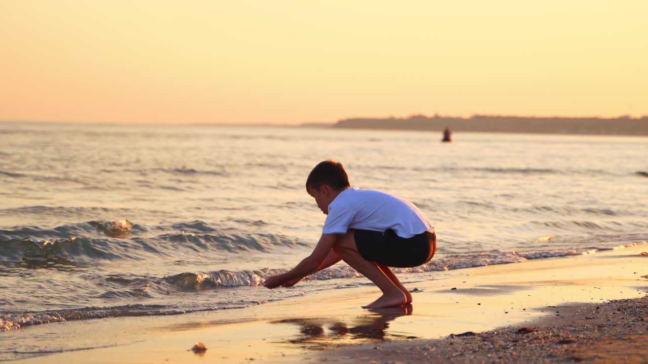 Boy plays in sea water in the evening. Side view of a boy in white t-shirt and shorts playing on sea shore at sunset.