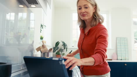 mature woman in kitchen at home cooking meal following recipe on digital tablet