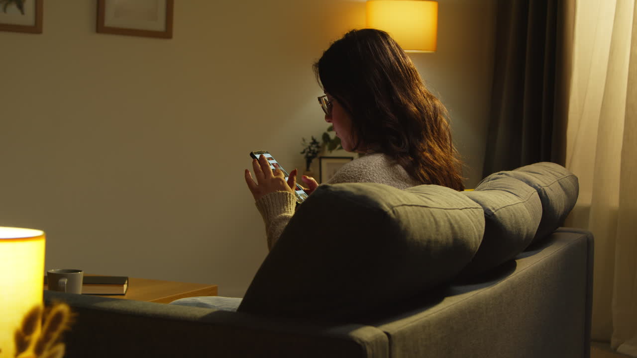 Rear View Of Young Woman At Home Sitting On Sofa With Mobile Phone Scrolling Through Internet In The Evening