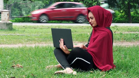 Happy-Afghan-woman-in-hijab-working-on-laptop-computer-on-green-lawn-near-busy-road