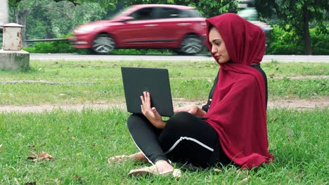 Glückliche-Afghanische-Frau-Im-Hijab,-Die-An-Einem-Laptop-Auf-Grünem-Rasen-In-Der-Nähe-Einer-Stark-Befahrenen-Straße-Arbeitet