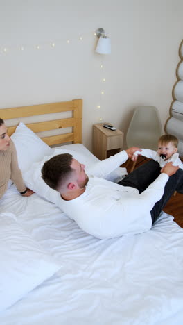 father holds his little baby on his leg waving him. dad and son are playing adorably. mother sits watching beside. top view. vertical video.