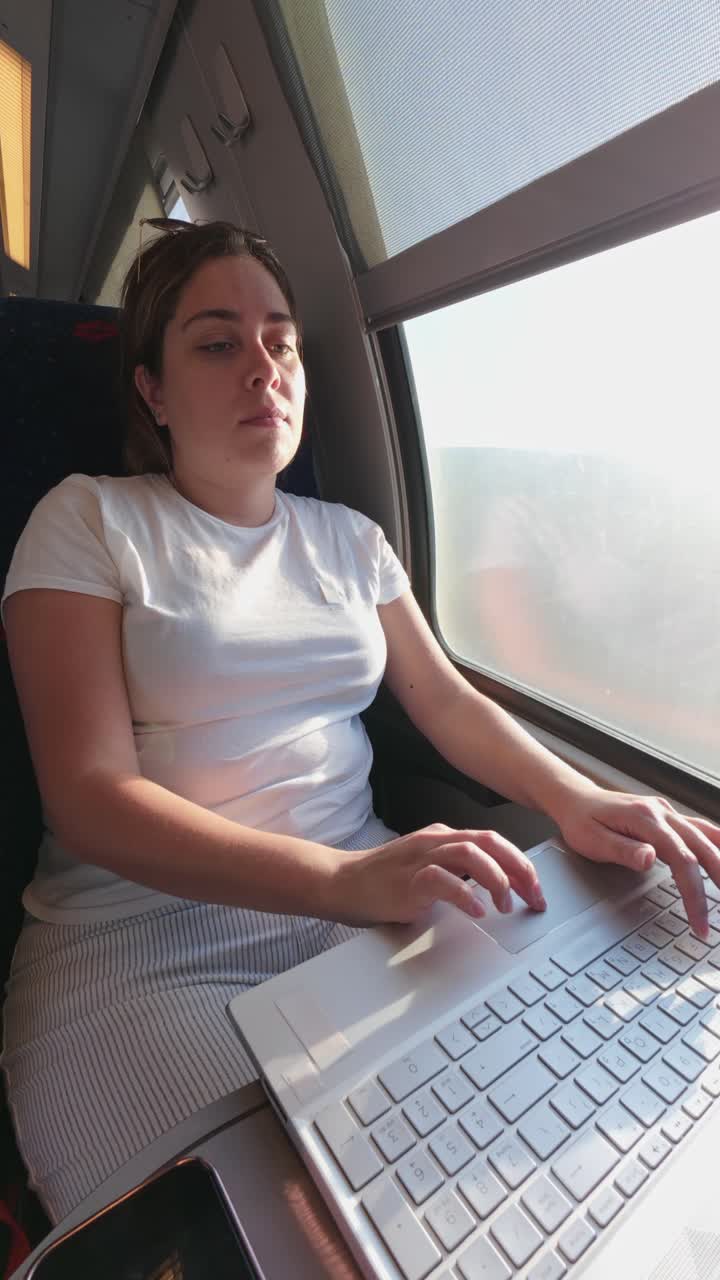 A young woman concentrates on her work, typing on a laptop computer while seated on a train