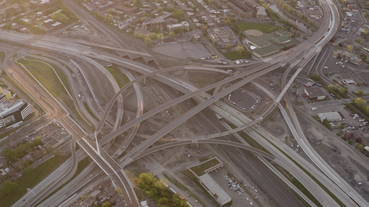Curving ramps of the Turcot Interchange cross rail yards and work zones in soft evening light as the camera glides overhead.