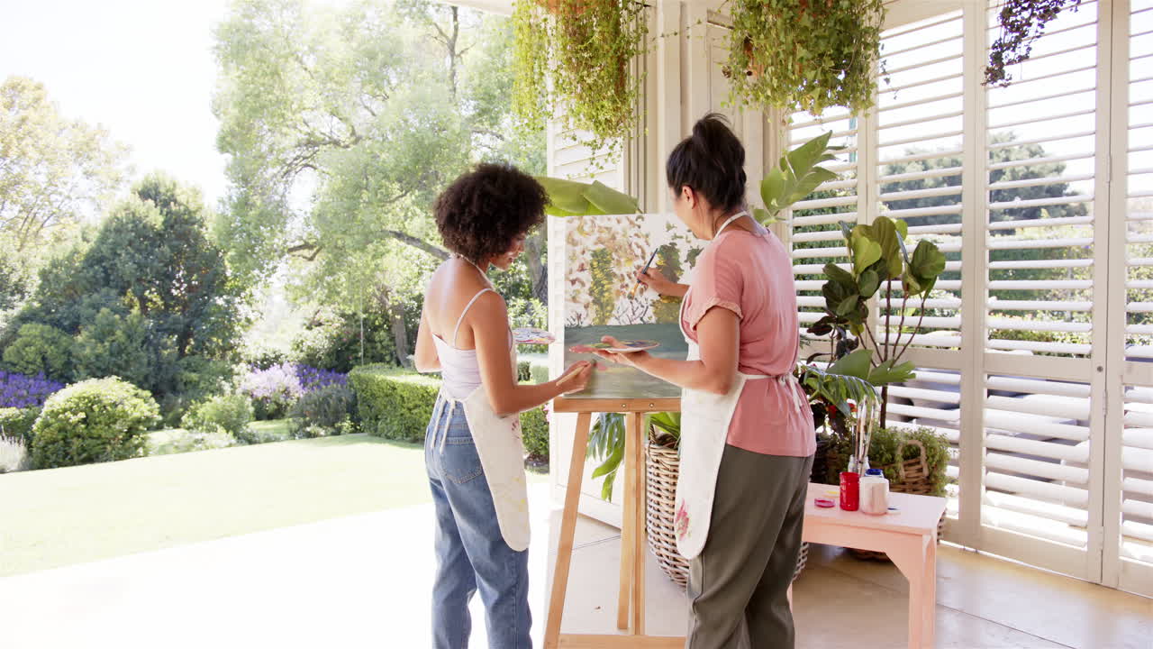 Painting on canvas, mother and adult daughter collaborating in art studio surrounded by plants