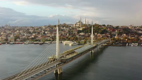 Drone-Aéreo-De-Una-Mañana-Nublada-De-Amanecer-En-Estambul,-Turquía,-Con-Vistas-A-Hagia-Sophia-En-Lo-Alto-De-Una-Colina-Y-Un-Puente-De-Metro-Halic-Mientras-Las-Gaviotas-Pasan-Volando-Y-Los-Barcos-Esperan-Para-Cruzar-El-Río-Bósforo