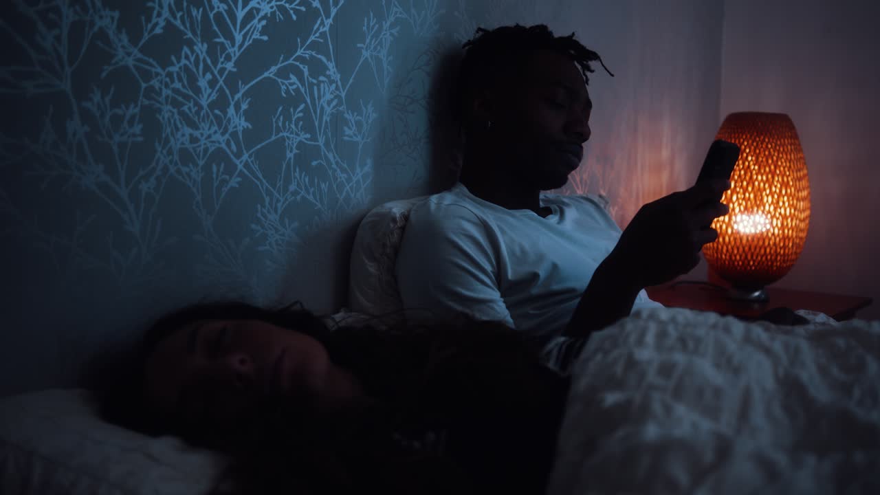 hombre frota los ojos y usa el teléfono inteligente mientras está sentado en la cama tarde en la noche