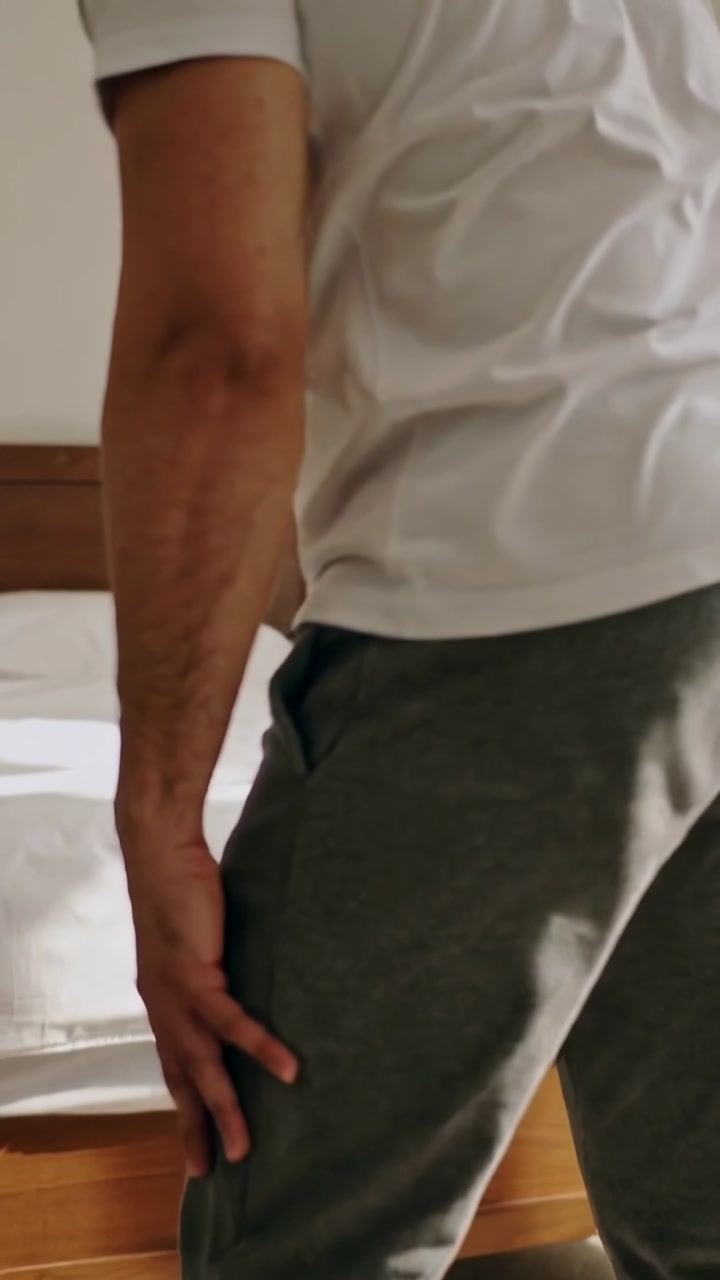 Vertical video: After leaning over bed, man adjusting gray sweatpants, preparing to rise in bedroom