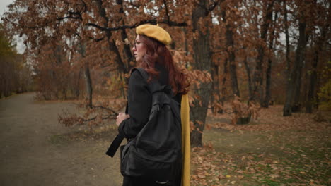 vista trasera de una mujer con muffler amarillo y boina caminando por el bosque, sosteniendo la correa de la bolsa y mirando hacia el lado, el cabello balanceándose en la brisa de otoño, la figura borrosa en el fondo