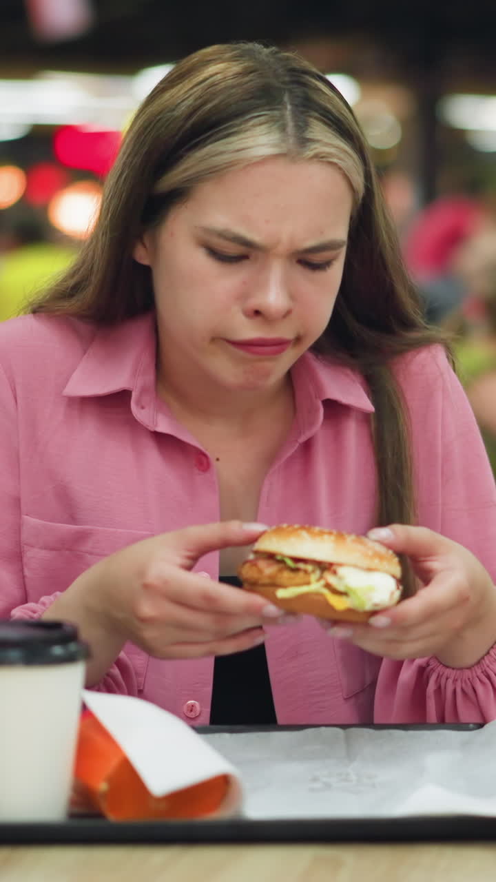 dama de vestido rosa huele hamburguesa con una expresión de descontento, luego lo deja caer, ella alcanza una servilleta para limpiarse la mano, mostrando asco, con vista borrosa de la gente comiendo en el fondo
