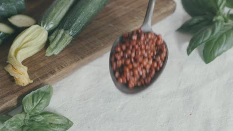 toma desde arriba de una mesa en la que se coloca una cuchara de plata llena de pimiento rojo, desde el voladizo los granos caen por todas partes, en la escena también hay calabacines y hojas de albahaca