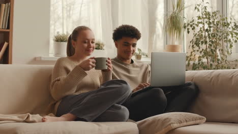 young couple relaxing on couch with laptop and drinks