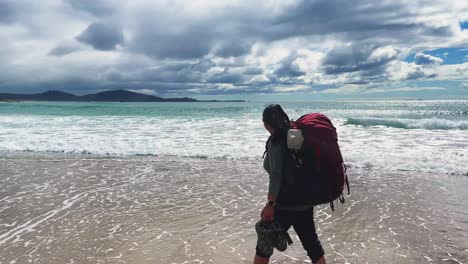 asian female backpacker walks in ocean waves at bright cloudy beach