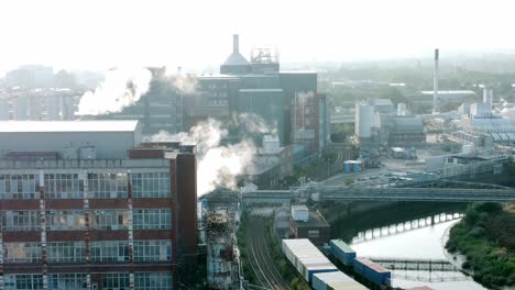 Industrielles-Chemisches-Lagergebäude-Am-Fluss-Mit-Fabrikdampfemissionen-Im-Sonnenlicht-Des-Frühen-Morgens,-Das-Aus-Der-Luft-Absteigt