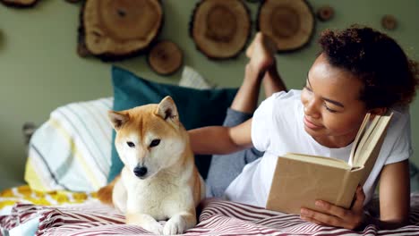 una joven afroamericana está leyendo un libro y acariciando a su perro mascota que descansa en la cama en un hermoso apartamento moderno. el animal está disfrutando del amor y el cuidado de su dueño.