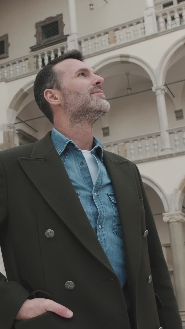 Uomo con cappotto e camicia di jeans che guarda in su in un cortile