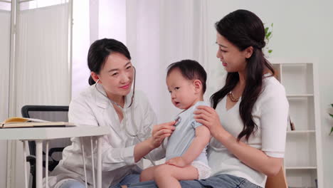 Expert examining cute little baby with stethoscope. Kid happily smiling and pointing on pediatrician. Mother holding her boy. Doctor visit his patient at clinic.