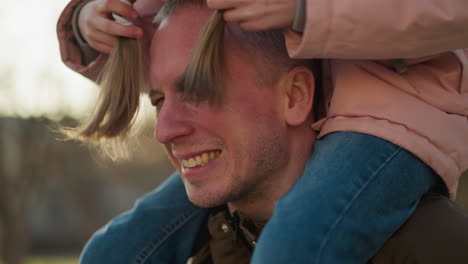 a little girl in a pink cap and jacket sits joyfully on her dad's neck as they walk through a sunlit park. she playfully bends her head down and uses her hair to play on his face