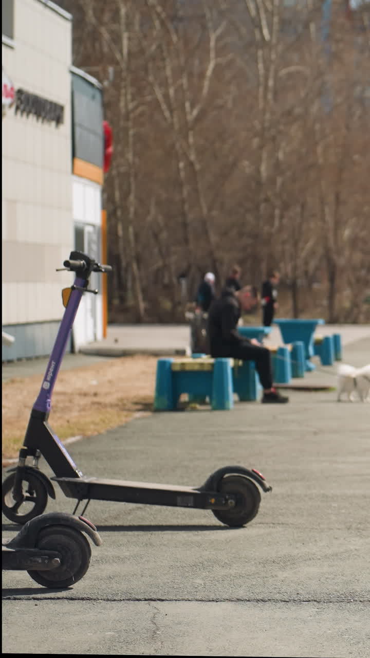 Close view of an urban parking lot featuring an electric scooter in the foreground, parked cars, and people relaxing on benches