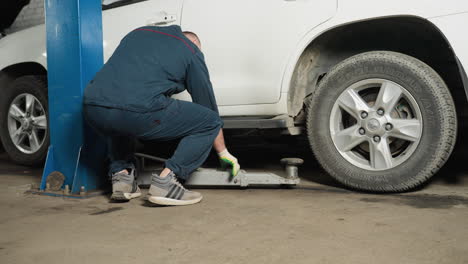 back view of a mechanic positioning a hydraulic jack under a white suv in a workshop, with a blue pole visible by the side, showing detailed car maintenance in progress