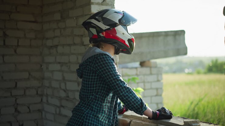 A lady stands contemplatively by an old building window, resting her hand on the ledge, her helmet visor is open as she gazes out into the peaceful landscape, seemingly lost in thought