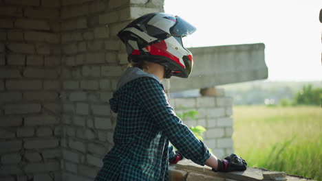 a lady stands contemplatively by an old building window, resting her hand on the ledge, her helmet visor is open as she gazes out into the peaceful landscape, seemingly lost in thought